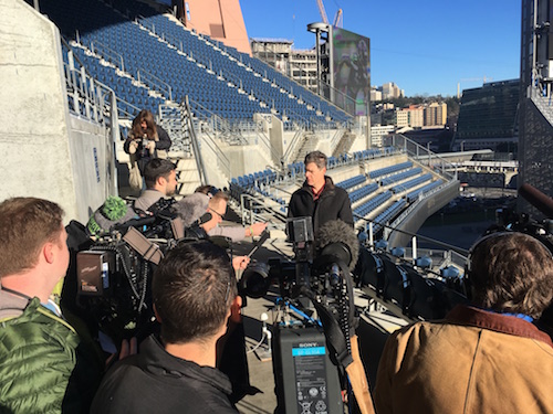 John Vidale stands in the stands of Century Link Field in front of several reporters holding cameras and microphones pointed at him.